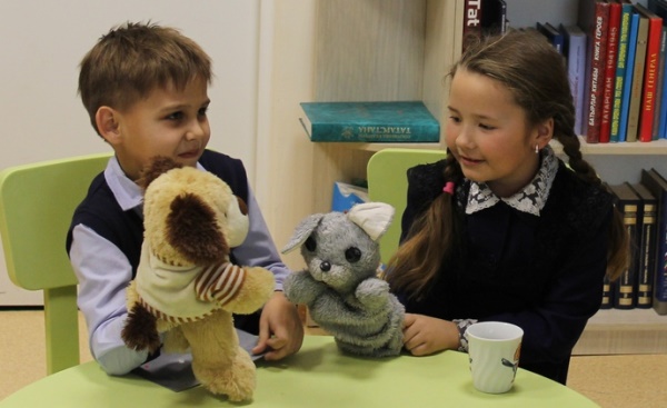 Мальчик и девочка сидят за столом с мягкими куклами - собакой и кошкой Мальчик и девочка сидят за столом с мягкими куклами - собакой и кошкой